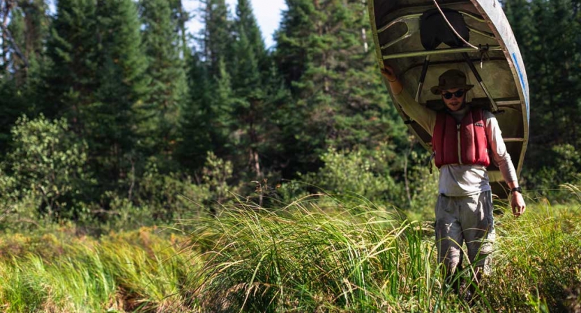 a young person wearing a life jacket carries a canoe on their shoulders through tall green grass